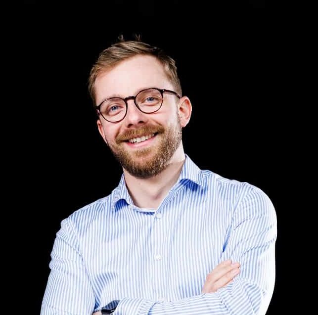 Muž s úsměvem, brýlemi a modrobílou košilí, stojící na černém pozadí, s rukama zkříženýma na hrudi.