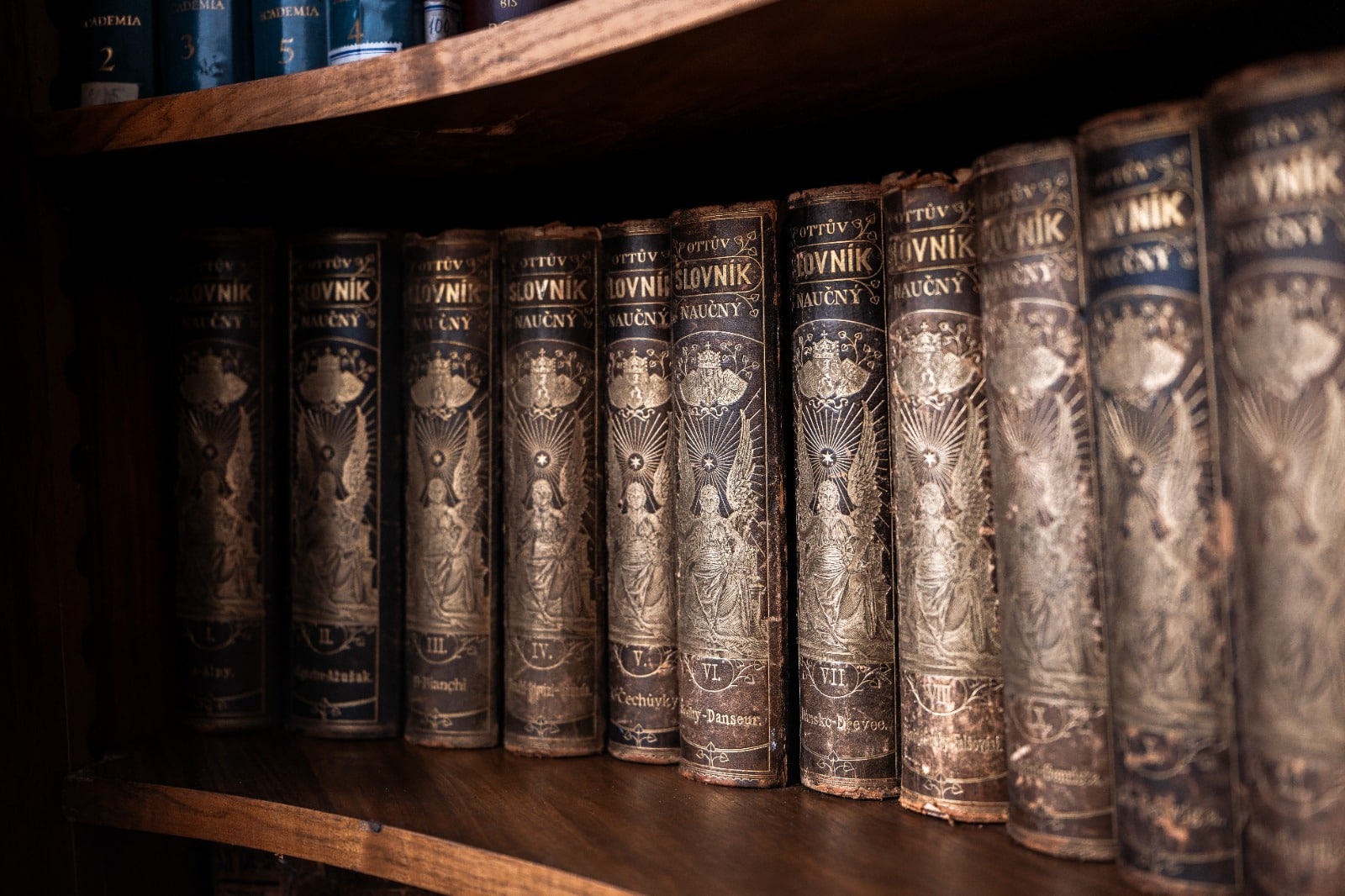 Stará knihovna s historickými svazky, Ottův slovník naučný, laděná do tmavých barev na dřevěných policích.