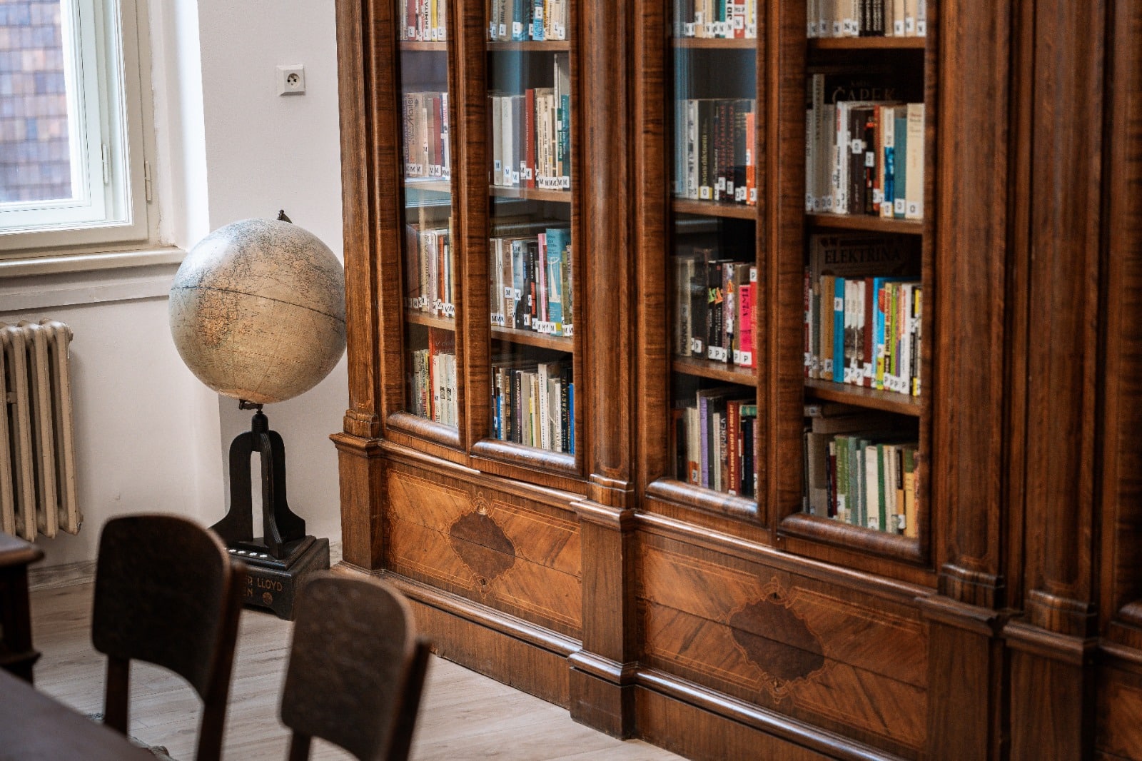 Starobylá knihovna s dřevěnými regály, plná knih, vedle glóbusu na stojanu, osvětlená denním světlem z okna.