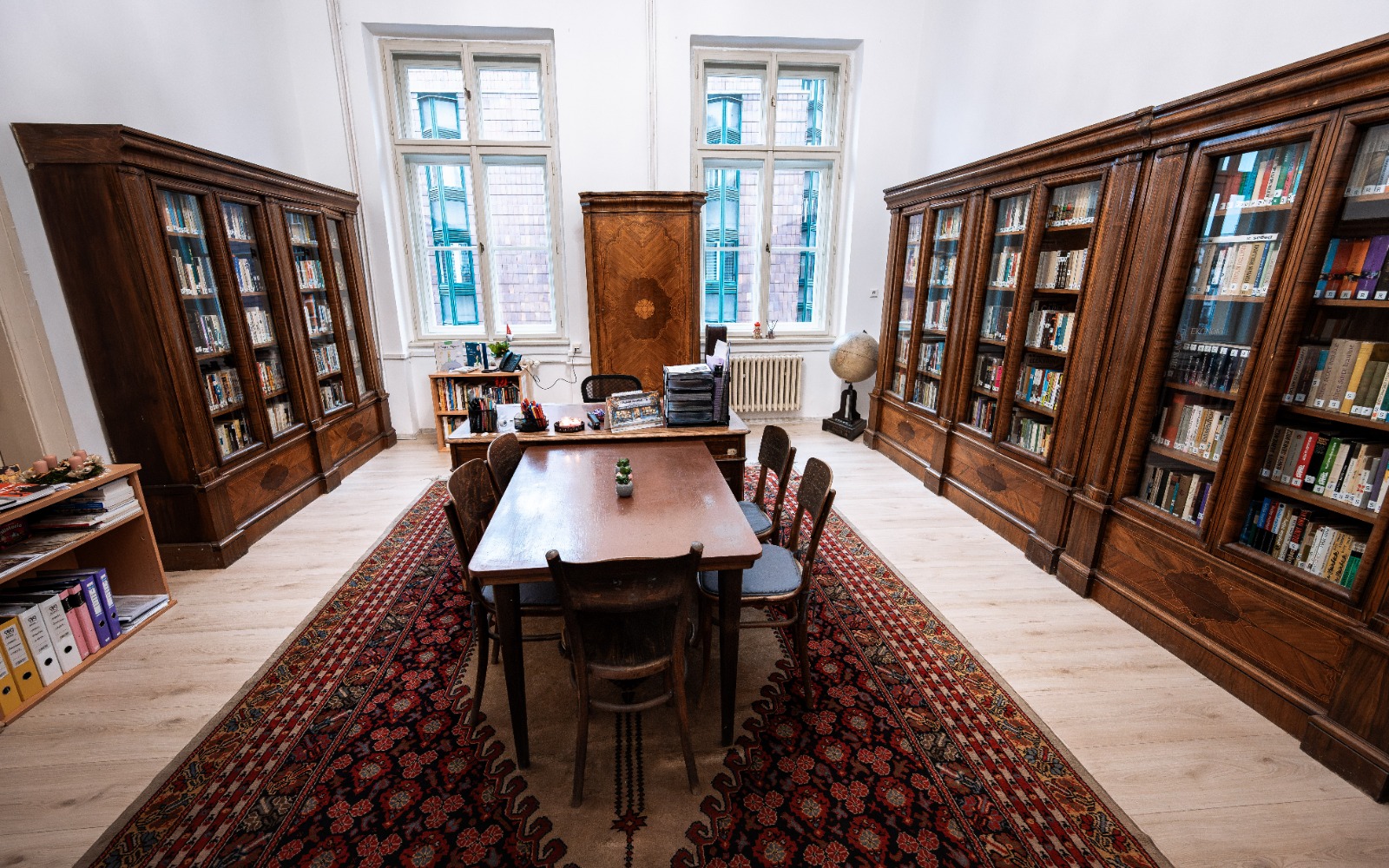 Historická knihovna s dřevěným nábytkem, kobercem a stolem obklopeným knihami pod velkými okny.