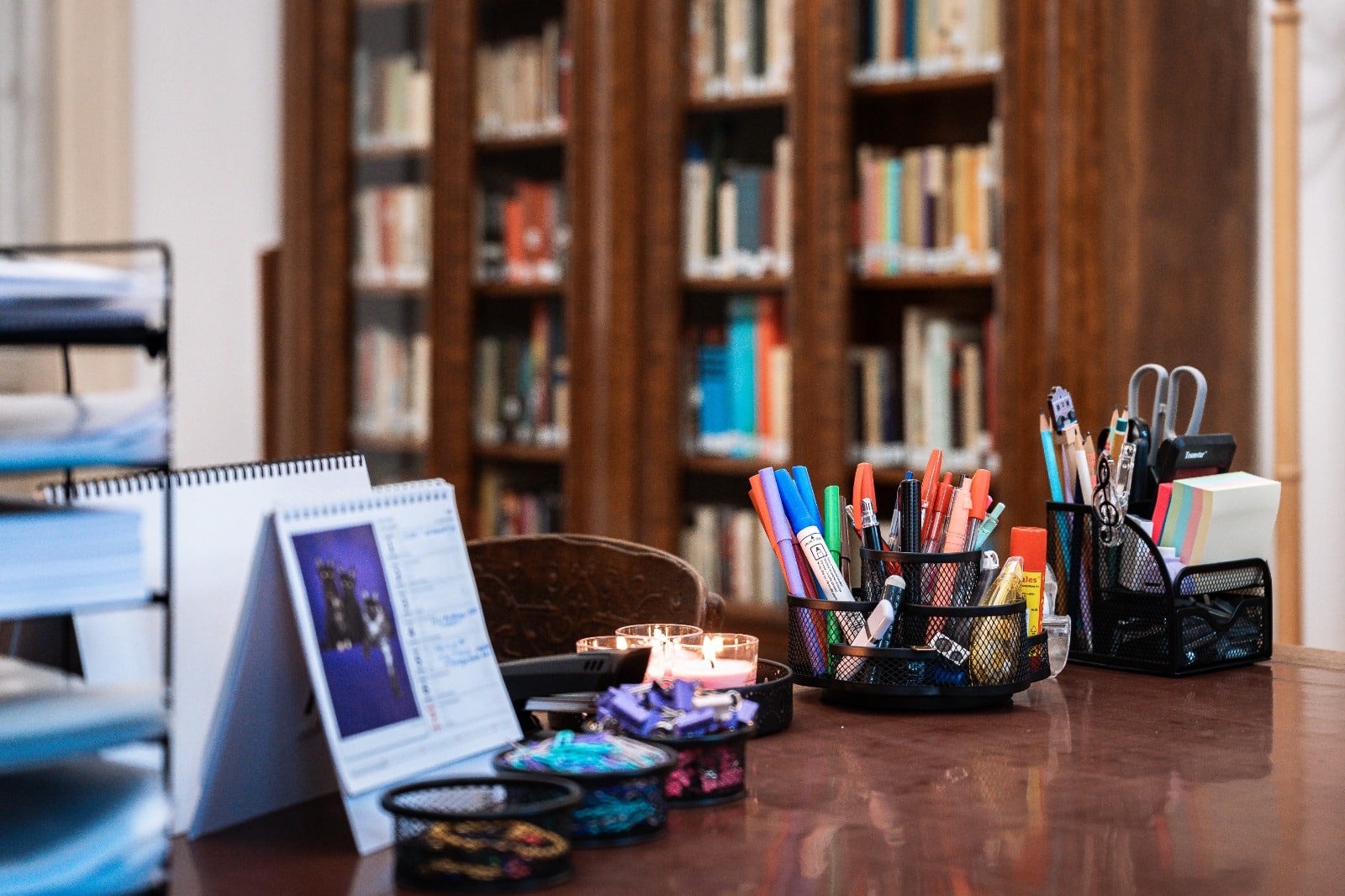 Pracovní stůl s kancelářskými potřebami před knihovnou plnou knih v útulném prostředí kanceláře.