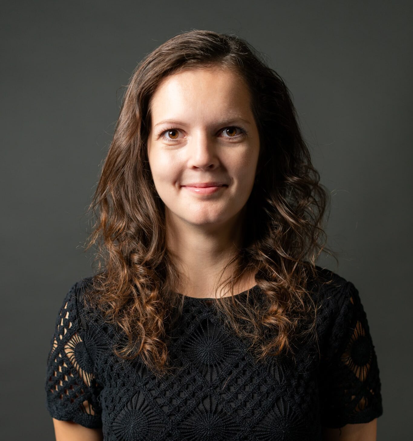 Žena s kudrnatými vlasy v černé krajkové halence pózuje na neutrálním pozadí, usmívá se přímo do kamery.