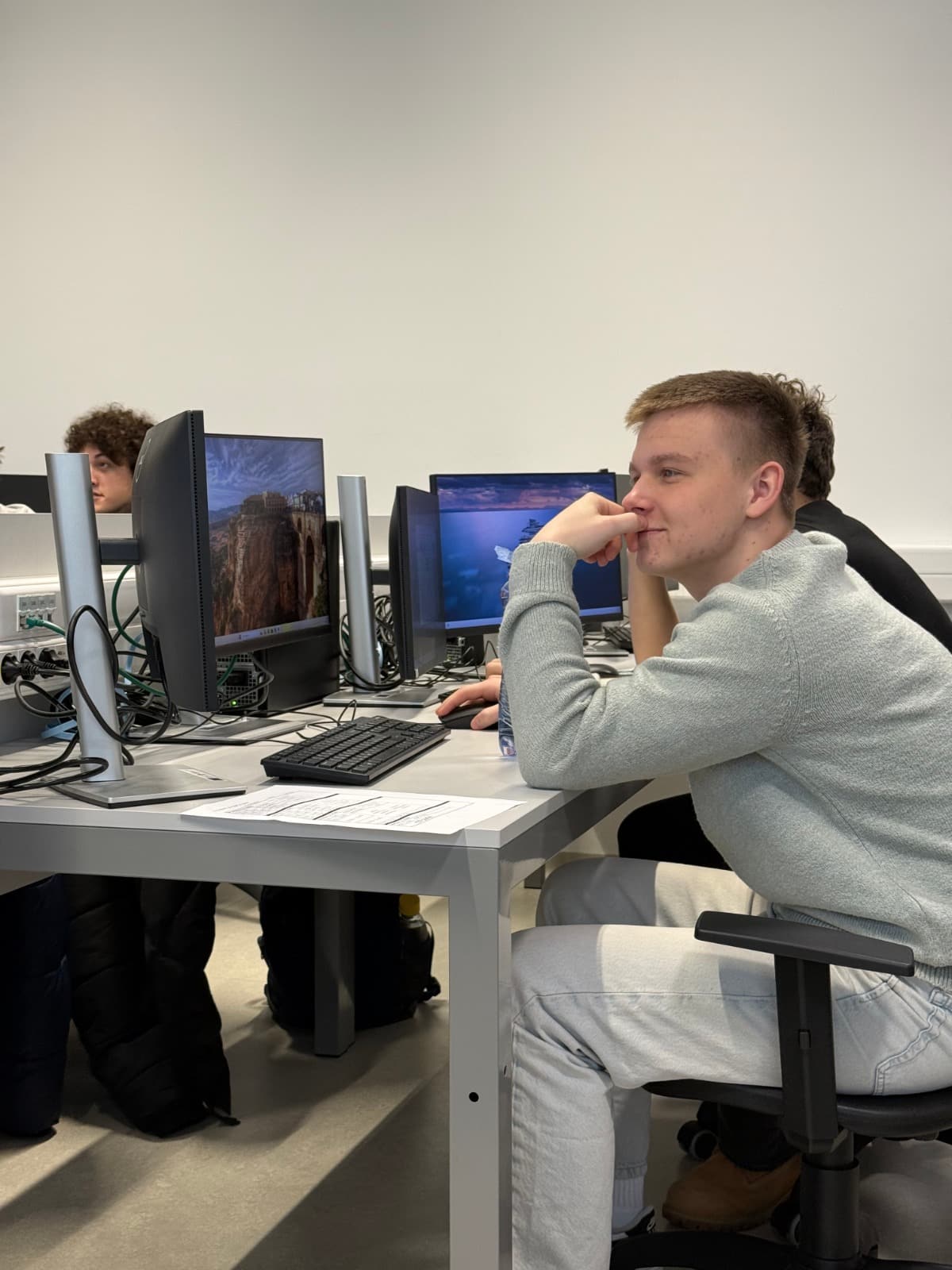 Student pracuje na počítači v učebně, zamyšleně sleduje obrazovku, vedle něj další studenti při výuce.